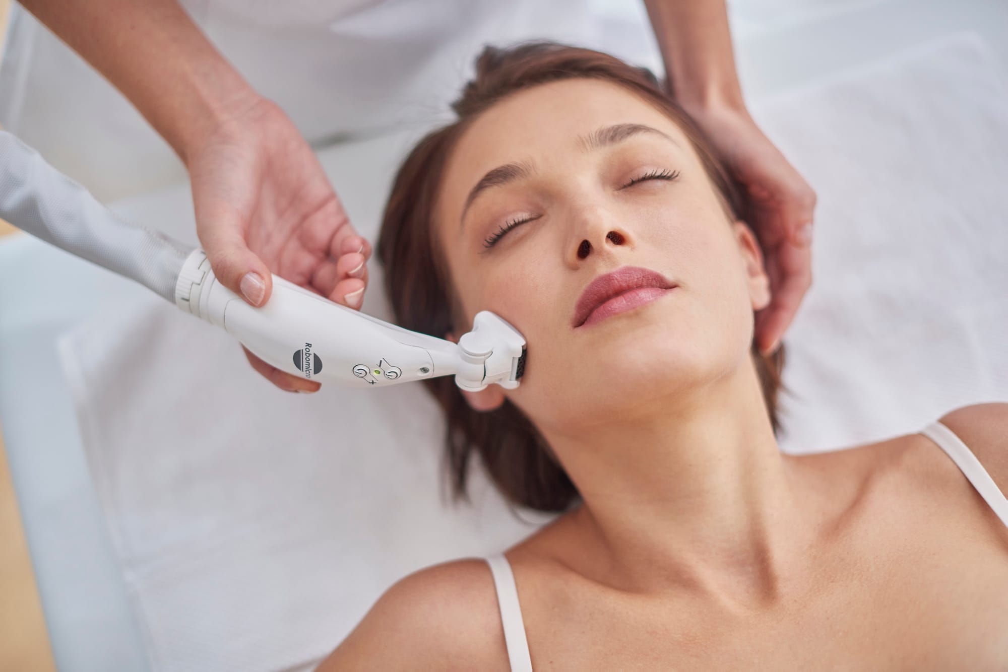 An ICOONE handpiece guided along a client&rsquo;s cheek as she rests with her eyes closed.